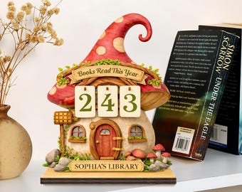 May include: A whimsical mushroom-shaped book counter with a red and white spotted cap. The counter displays the number "243" on hanging tags. The base reads "Sophia's Library". Decorative mushrooms and foliage surround the structure. Books are visible in the background.