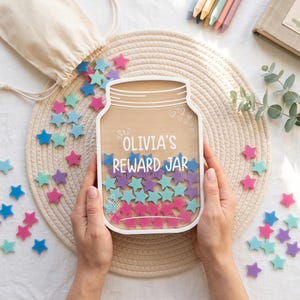 Könnte beinhalten: Ein Belohnungsglas aus Holz mit einer transparenten Vorderseite und der weißen Aufschrift OLIVIA'S REWARD JAR. Das Glas ist mit kleinen, bunten sternförmigen Plättchen in Rosa, Lila und Blau gefüllt, weitere Sterne liegen verstreut daneben.