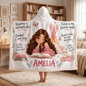 May include: A white hooded blanket featuring a graphic of a person with long brown hair reading a book. Text includes Reading is my happy place, Booked and cozy, A little library in my heart, Fiction is my favorite escape, Readers make the best dreamers, Coffee. Books. Repeat. and the name AMELIA, decorated with butterflies, flowers, and hearts.