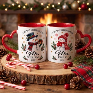 May include: Two white ceramic mugs with red interiors and heart-shaped handles. Each mug features a snowman illustration, pine branches, and gold stars. Text reads: OUR FIRST CHRISTMAS AS Mr. WILLIAM 2026 and OUR FIRST CHRISTMAS AS Mrs. EMILY 2026.