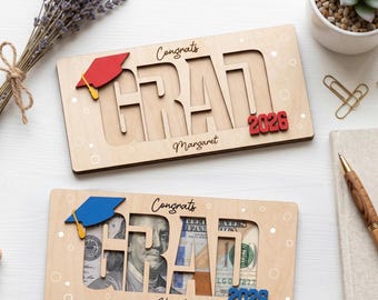 May include: Two wooden graduation gift boxes with the word "GRAD" cut out, with a graduation cap and the year "2026". One box has the name "Margaret" and a red cap, the other has the name "Edward" and a blue cap. The boxes are designed to hold cash.