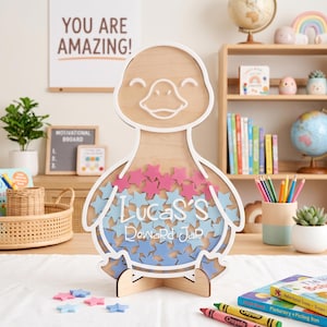 May include: A light-colored wooden reward jar shaped like a duck, featuring a white outline and a clear front panel filled with small pink and blue wooden stars. The text "Lucas's Reward Jar" is printed in white across the center of the duck's body.