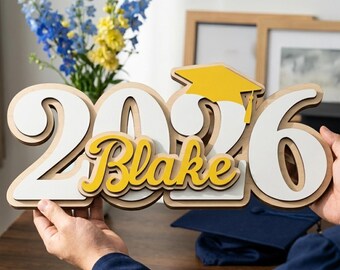 May include: A wooden graduation decoration with the year 2026 in white and a yellow graduation cap. The name "Blake" is in yellow script. The decoration is held up by hands, with a graduation cap in the background.