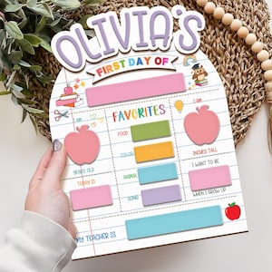May include: A wooden first day of school sign with the name "Olivia's" in purple and white. The sign has sections for favorites, including food, color, and animal, and a section for inches tall. The sign is held by a person.