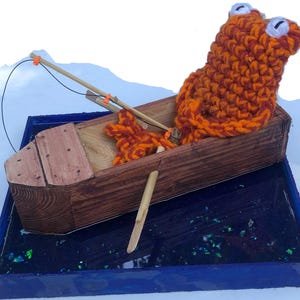 May include: A handcrafted wooden boat with a crocheted orange and brown frog figure. The boat has a fishing rod and sits in a blue resin base with iridescent accents. The frog has white eyes.