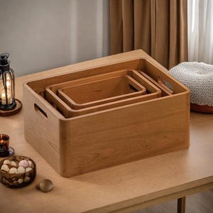 Könnte beinhalten: Satz von drei Holz-Nesting-Boxen mit abgerundeten Ecken und integrierten Griffen. Die Boxen sind aus hellem Holz gefertigt und auf einem Holztisch ineinander angeordnet. Die größte Box ist etwa 46 cm lang.