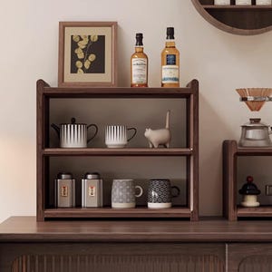 May include: A dark wood shelf unit displaying various items. The top shelf holds two bottles and a framed picture. The middle shelf features a teapot, a mug, and a cat figurine. The bottom shelf has two canisters and two mugs. The items are arranged on a dark wood surface.