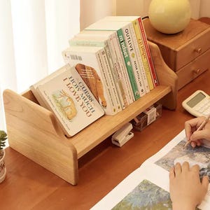 May include: A wooden desk organizer with two shelves holding books. The top shelf displays books with titles like "OUTLIVE" and "What If?". The bottom shelf holds a book titled "Guess How Much I Love You". The organizer is made of light-colored wood.