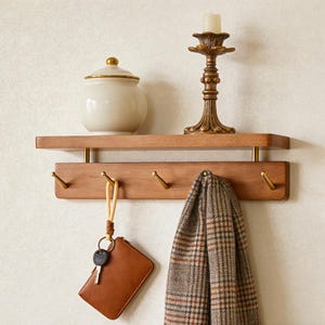 May include: A wooden wall shelf with a lower row of hooks. A tan scarf, a brown leather wallet with keys, and a cream-colored jar with a gold lid hang from the shelf. A gold candlestick with a white candle sits on the shelf.