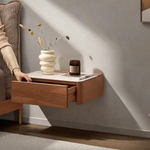 Może przedstawiać: Pływający drewniany stolik nocny z otwartą szufladą. Stolik nocny ma biały blat i jest ozdobiony kremowym wazonem z suszonymi kwiatami, małym słoiczkiem i książką. Stolik nocny jest zamontowany na szarej ścianie.