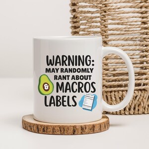 May include: White ceramic mug with a handle, featuring the text "WARNING: MAY RANDOMLY RANT ABOUT MACROS LABELS." An avocado graphic and a nutrition facts label are also on the mug. The mug sits on a small, round wooden coaster.