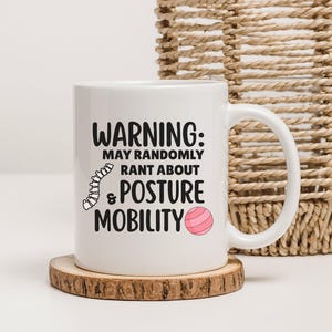 May include: White ceramic mug with the text "WARNING: MAY RANDOMLY RANT ABOUT & POSTURE MOBILITY" in black. The mug features a spine illustration and a pink exercise ball graphic. The mug sits on a wooden coaster.