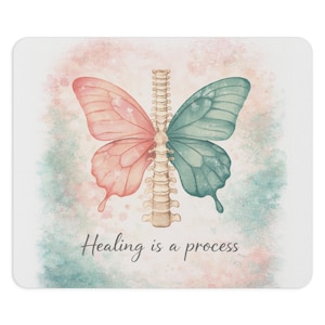 Puede incluir: Una alfombrilla de ratón con una ilustración de acuarela de una mariposa, un ala rosa y otra turquesa, dividida por una columna vertebral. El texto "Healing is a process" está escrito debajo de la mariposa.