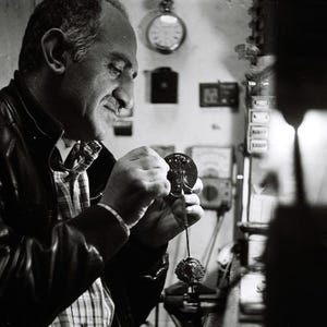 Puede incluir: Un hombre con una chaqueta de cuero negra y una camisa con estampado blanco y negro está trabajando en un pequeño objeto redondo de plata. Está sujetando el objeto con la mano izquierda y usando la derecha para ajustarlo. El objeto parece ser un reloj o un reloj.