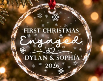 Può includere: Una decorazione natalizia circolare in vetro trasparente sfaccettato, appesa a un nastro rosso. Presenta la scritta bianca "FIRST CHRISTMAS Engaged DYLAN & SOPHIA 2026" con grafiche di fiocchi di neve e cuori.