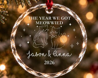 Può includere: Un ornamento in vetro trasparente, circolare e sfaccettato, appeso a un nastro rosso. Presenta una scritta bianca incisa che recita THE YEAR WE GOT MEOWWIED, Jason & Linna, 2026, con il disegno di due gatti, uno con velo e uno con papillon.