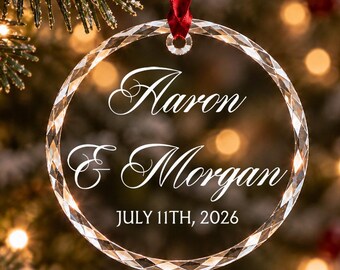 Può includere: Un ornamento circolare in vetro trasparente con bordo sfaccettato, appeso a un nastro rosso. Il centro è inciso con i nomi Aaron & Morgan e la data JULY 11TH, 2026 in caratteri bianchi corsivi e stampatello sulla superficie del vetro.
