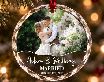 Può includere: Un ornamento circolare in vetro sfaccettato con una fotografia di una coppia che si abbraccia all'aperto. Il testo recita Adam & Brittany MARRIED AUGUST 1ST, 2026, con un disegno a linee bianche di due cuori intrecciati sopra i nomi.