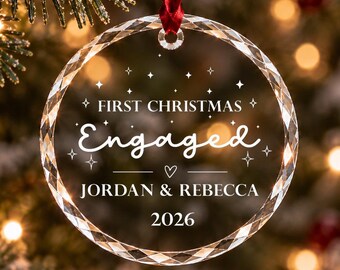 Può includere: Un addobbo natalizio circolare in vetro trasparente con bordo sfaccettato, appeso a un nastro rosso. L'ornamento è inciso con il testo "FIRST CHRISTMAS Engaged", "JORDAN & REBECCA" e "2026", decorato con piccoli simboli di stelle e cuori.