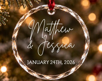 Può includere: Un ornamento circolare in vetro trasparente con bordo sfaccettato, appeso a un nastro rosso. L'ornamento presenta il testo stampato in bianco Matthew & Jessica e la data JANUARY 24TH, 2026, centrati sulla superficie trasparente.