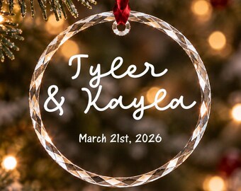 Può includere: Un ornamento in vetro trasparente, circolare e sfaccettato, appeso a un nastro rosso. Al centro presenta il testo bianco "Tyler & Kayla" in carattere corsivo, seguito dalla data "March 21st, 2026" in un carattere semplice e senza grazie.