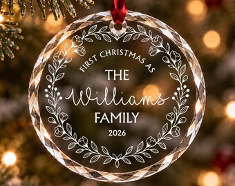 Può includere: Un addobbo natalizio circolare in vetro trasparente con bordo sfaccettato, appeso a un nastro rosso. Il centro presenta un design a ghirlanda floreale inciso con il testo "FIRST CHRISTMAS AS THE Williams FAMILY 2026" in bianco.