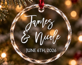 Può includere: Un ornamento circolare in vetro trasparente con bordo sfaccettato, appeso a un nastro rosso. Il centro presenta il testo stampato in bianco James & Nicole e la data JUNE 6TH, 2026 in una combinazione di caratteri corsivi e sans-serif.