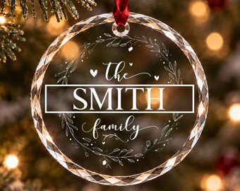 Può includere: Un ornamento circolare in vetro trasparente sfaccettato appeso a un nastro rosso. Il centro presenta il testo 'the SMITH family' racchiuso in un rettangolo orizzontale, circondato da un delicato disegno a ghirlanda con cuori.