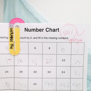 May include: A number chart with a pink and yellow paper clip that says "Mrs. Robinson" on it. The chart is marked with a 100% and "great job!" in red. The chart is counting by 2.