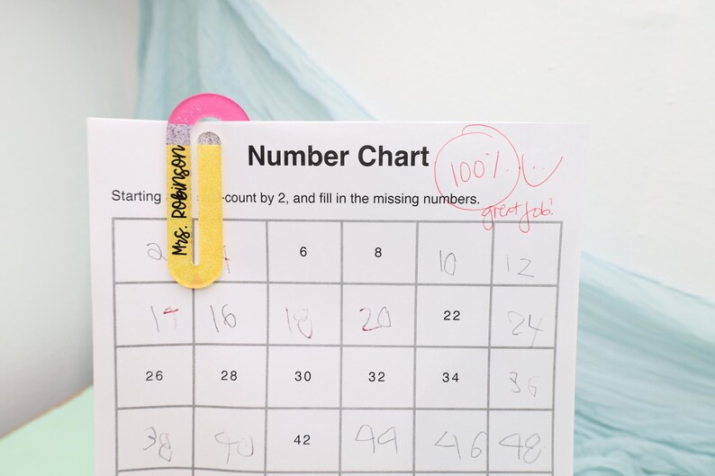 May include: A number chart with a pink and yellow paper clip that says "Mrs. Robinson" on it. The chart is marked with a 100% and "great job!" in red. The chart is counting by 2.
