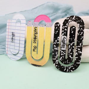 May include: Three decorative paperclip-style bookmarks. One is designed to look like a pencil, another has a notebook paper design with the name "George", and the third has a black and white pattern with the name "Taylor".