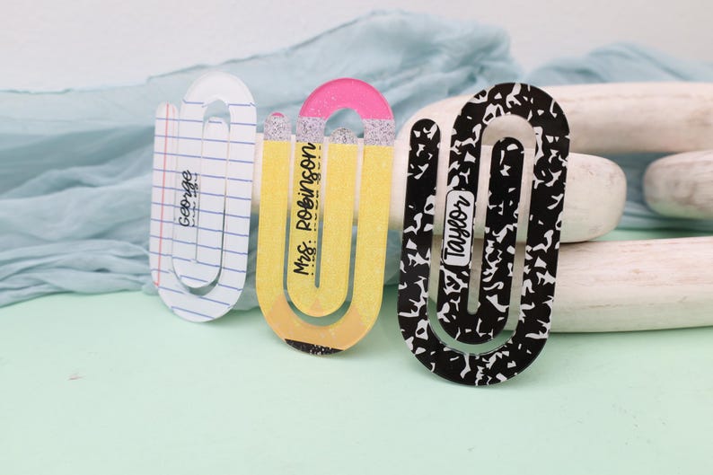May include: Three decorative paperclip-style bookmarks. One is designed to look like a pencil, another has a notebook paper design with the name "George", and the third has a black and white pattern with the name "Taylor".