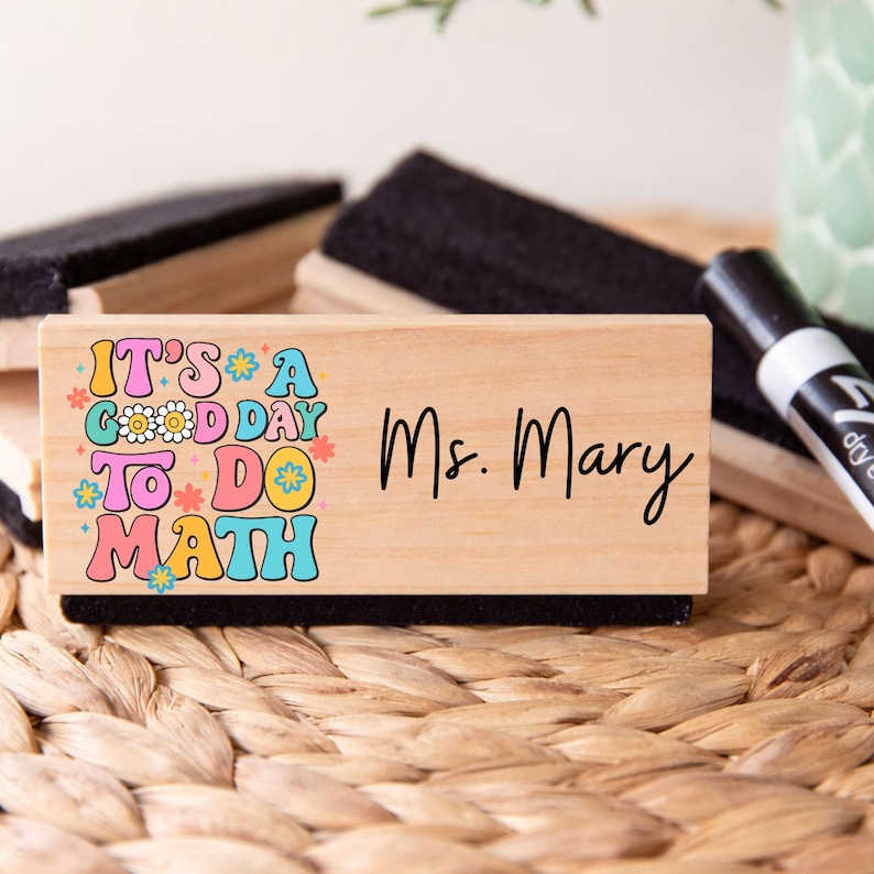 Puede incluir: Un letrero de escritorio de madera con el texto "It's a good day to do math" en una fuente retro colorida. El nombre "Ms. Mary" est&aacute; escrito en escritura negra. Una goma de borrar negra est&aacute; adjunta en la parte inferior.