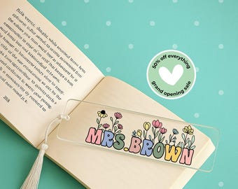 Può includere: Segnalibro trasparente in acrilico con il nome "MRS. BROWN" in lettere colorate in stile retrò, decorato con illustrazioni floreali. Il segnalibro è appoggiato su un libro aperto con un nappa, su sfondo turchese.