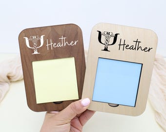 Può includere: Due portamemo in legno, uno marrone scuro e uno marrone chiaro, ciascuno con un'apertura rettangolare e il nome "Heather" e un simbolo medico. Uno ha un post-it giallo, l'altro blu.