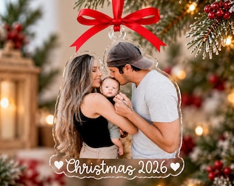 Può includere: Una decorazione natalizia in acrilico trasparente a forma di campana con la fotografia stampata di una famiglia di tre persone. L'ornamento è sormontato da un fiocco rosso e presenta la scritta bianca "Christmas 2026" tra due piccoli cuori.