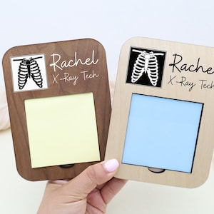 May include: Two wooden memo holders, one dark brown and one light brown, each with a square cutout for a notepad. The top of each holder features a black and white rib cage graphic and the text "Rachel X-Ray Tech."