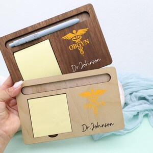 May include: Two wooden desk organizers, one light and one dark, each with a slot for a pen, a notepad, and a medical symbol. The dark organizer has the text "OBGYN" and "Dr. Johnson". The light organizer also has the text "Dr. Johnson".