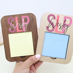 May include: Two wooden memo holders, one dark brown and one light brown, each with a rectangular opening for a notepad. The word "SLP" and the name "Wesley" are printed on the top in pink and black.