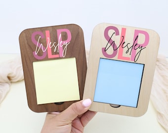 Op de afbeelding: Twee houten memohouders, één donkerbruin en één lichtbruin, elk met een rechthoekige opening voor een notitieblok. Het woord "SLP" en de naam "Wesley" zijn in roze en zwart gedrukt.