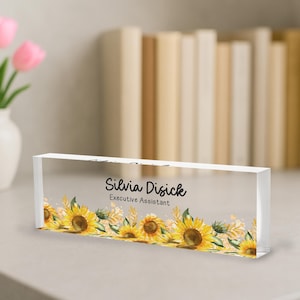 May include: A clear acrylic desk nameplate featuring the name "Silvia Disick" and the title "Executive Assistant" in black script. The bottom edge is adorned with a watercolor design of yellow sunflowers and green leaves.