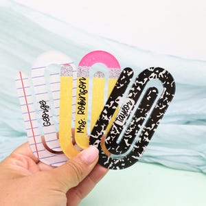 May include: A collection of decorative paper clips in the shape of pencils and notebook paper. The clips are in various colors, including yellow, pink, and black and white. Each clip has a name printed on it: George, Mrs. Robinson, and Taylor.