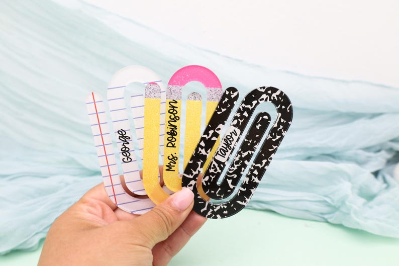 May include: A collection of decorative paper clips in the shape of pencils and notebook paper. The clips are in various colors, including yellow, pink, and black and white. Each clip has a name printed on it: George, Mrs. Robinson, and Taylor.