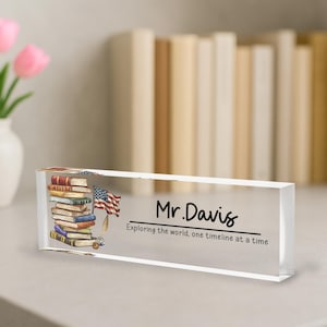 May include: A rectangular clear acrylic desk nameplate with the name "Mr. Davis" in black script. The nameplate features an illustration of a stack of books, an American flag, and the text "Exploring the world, one timeline at a time."