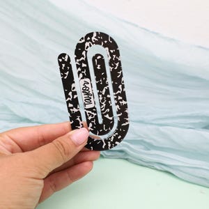 May include: A black and white composition notebook-themed bookmark. The bookmark is held in a hand and features the name "Taylor" in a white font. The background is a light blue fabric.