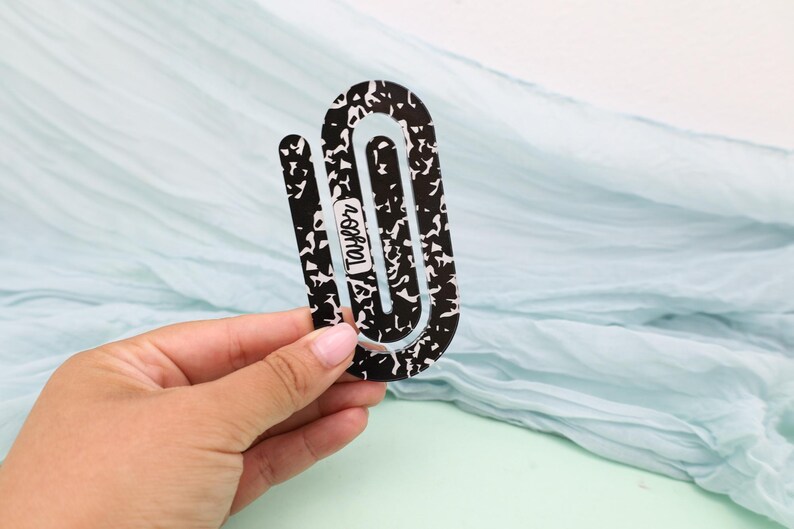 May include: A black and white composition notebook-themed bookmark. The bookmark is held in a hand and features the name "Taylor" in a white font. The background is a light blue fabric.