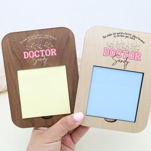 May include: Two wooden memo holders, one dark brown and one light brown, each with a rectangular opening for a notepad. The text "DOCTOR Sandy" is printed in pink. The top of each holder has the text "You make the world a better place because of the lives you touch."