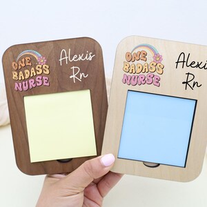 May include: Two wooden memo holders, one dark brown and one light brown, each with a rectangular opening. The text "ONE BADASS NURSE" is above the opening, with the name "Alexis Rn" below. One has a yellow pad, the other a blue pad.