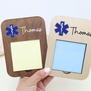 May include: Two wooden memo holders, one dark brown and one light brown, each with a blue medical symbol and the name "Thomas" in script. The dark brown holder has a yellow note, the light brown has a blue note.