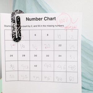 May include: A handwritten number chart, filled in with numbers counting by two. The chart is held by a black and white patterned paper clip. The chart has a grade of 100% and the words "great job" written in red.